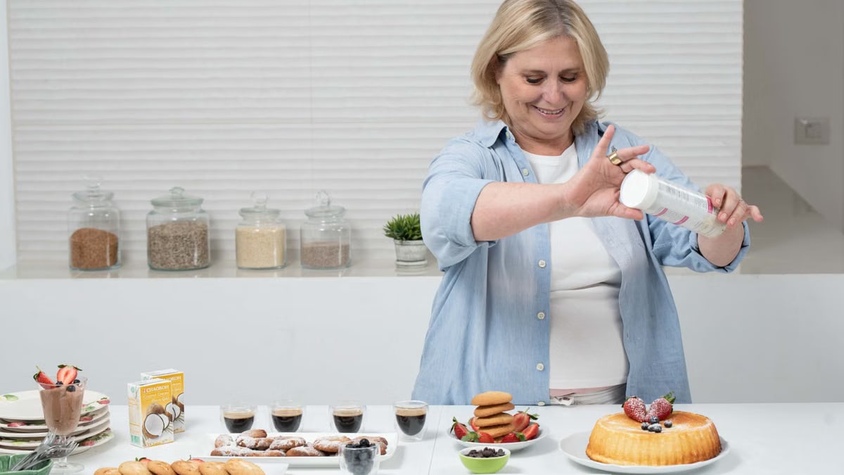 Smiling woman decorates cake with icing in a bright kitchen, surrounded by baked goods.